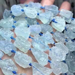 Könnte beinhalten: Eine Strähne aus hellblauen Aquamarin-Edelsteinkugeln, jede Kugel hat eine raue, unregelmäßige Form mit einem durchscheinenden, leicht trüben Aussehen. Die Kugeln sind auf einer blauen Schnur aufgefädelt.