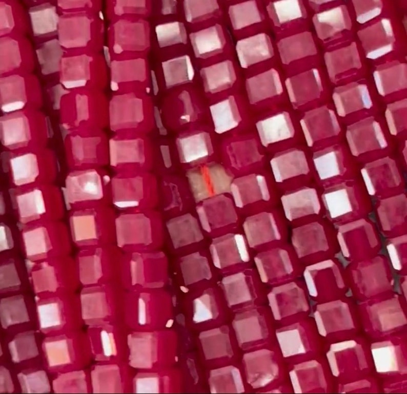 May include: Close-up of a strand of small, square, red glass beads. The beads have a slight iridescent sheen.