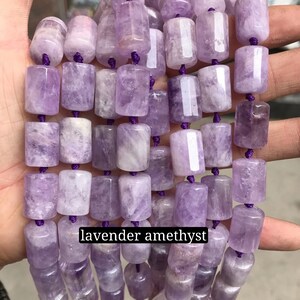 May include: A strand of lavender amethyst gemstone beads. The beads are cylindrical and have a smooth, polished finish. The beads are strung on a purple cord.