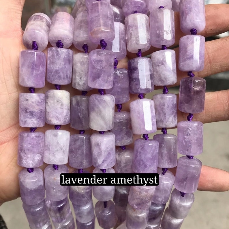 May include: A strand of lavender amethyst gemstone beads. The beads are cylindrical and have a smooth, polished finish. The beads are strung on a purple cord.