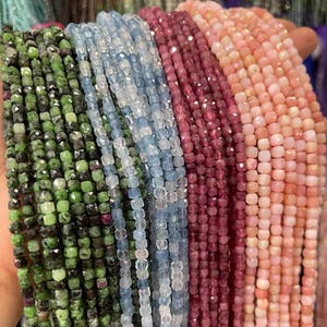 Pode incluir: Fios de contas de pedras preciosas em várias cores, incluindo verde, azul, vermelho e rosa. As contas são pequenas, de corte quadrado e enfiadas, criando uma exibição texturizada e colorida. Ideal para a criação de joias.