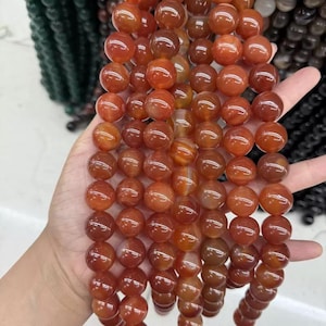 Puede incluir: Cadenas de cuentas redondas de piedras preciosas de color naranja rojizo pulidas. Las cuentas tienen una calidad translúcida con variaciones naturales en color y patrón. Las cuentas se sostienen en una mano, con otras cadenas de cuentas visibles en el fondo.