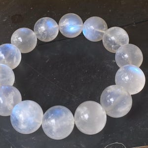 May include: A bracelet made of round, translucent moonstone beads. The beads have a subtle blue iridescence and are strung together to form a circular shape. The bracelet is displayed on a dark surface.