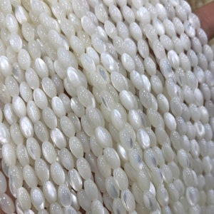 May include: A strand of white oval-shaped shell beads. The beads have a pearlescent sheen and are strung on a clear thread.