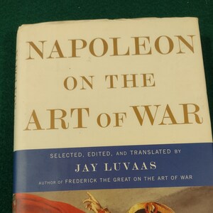 May include: A book titled "Napoleon on the Art of War" by Jay Luvaas. The cover features a painting of Napoleon Bonaparte on horseback, wearing a red coat and a bicorne hat.