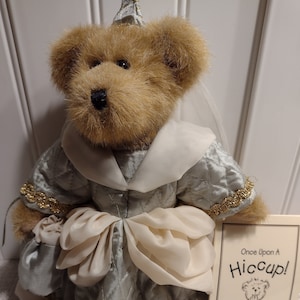 May include: A plush brown bear dressed as a princess in a light blue and white dress with a gold trim. The bear is wearing a crown and holding a white bag. The bear is standing on a white surface with a tag that says "Once Upon A Hiccup!" with a cartoon bear on it.