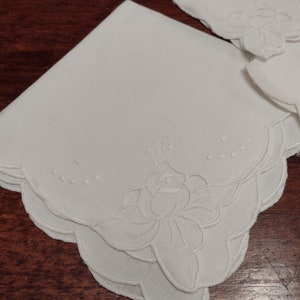 May include: Two white linen handkerchiefs with scalloped edges and embroidered floral designs. The handkerchiefs are folded and stacked on top of each other.
