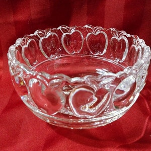 May include: A clear glass bowl with a scalloped rim and an apple design. The bowl is empty and sitting on a red tablecloth.