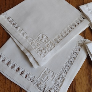 May include: Two white linen napkins with delicate lace trim and embroidered floral designs. The napkins are folded and stacked on a wooden surface.