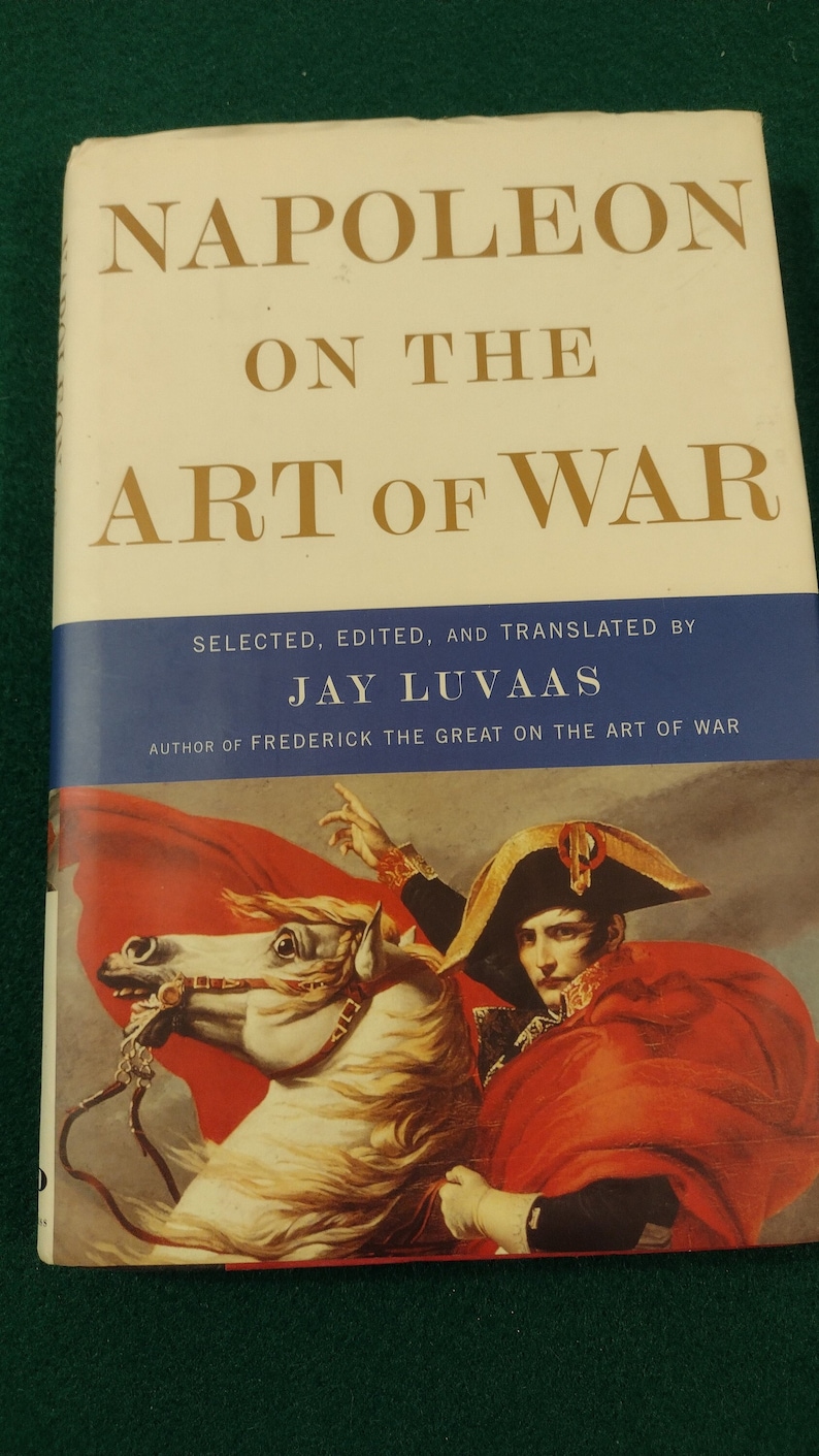 May include: A book titled "Napoleon on the Art of War" by Jay Luvaas. The cover features a painting of Napoleon Bonaparte on horseback, wearing a red coat and a bicorne hat.