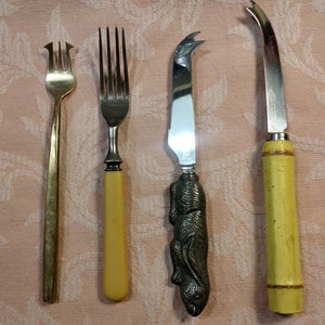 May include: Four utensils, including a gold fork, a yellow handled fork, a cheese knife with a rabbit handle, and a yellow handled cheese knife.