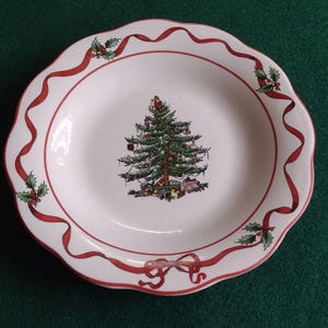 Pode incluir: Prato de Natal em cerâmica branca com borda ondulada e design de fita vermelha. O prato apresenta uma árvore de Natal decorada no centro e detalhes de azevinho. Um prato festivo para servir comida.