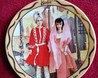 May include: A round, cream-colored ceramic plate with a gold rim. The plate features a colorful illustration of a man and woman in traditional Middle Eastern clothing. The text "Barbie" and "A Thousand and One Nights" are printed on the plate.