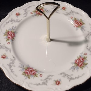 Puede incluir: Una fuente de dos niveles con asa dorada. El plato de porcelana blanca presenta un estampado floral de rosas rosas y detalles azules, con un diseño de volutas grises en los bordes. El borde está adornado con oro.