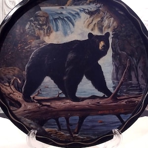 May include: A decorative black serving tray featuring a painting of a black bear walking on a log near a waterfall. The bear is the focal point, with a detailed depiction of its fur and expression. The tray has a scalloped edge and is supported by a clear plastic stand.