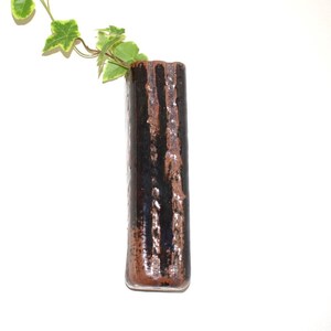 May include: A tall, rectangular ceramic vase with a dark brown and black glazed finish featuring vertical textured ridges. A small sprig of green and cream variegated ivy leaves extends from the top opening of the vase against a plain background.