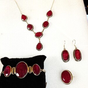 May include: A jewelry set featuring a necklace, earrings, bracelet, and ring. The set includes oval and teardrop-shaped, deep red gemstones set in silver-toned metal. The bracelet is displayed on a black velvet surface.