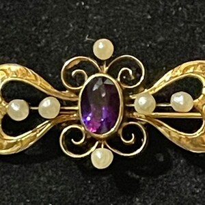 May include: A gold brooch with a purple amethyst gemstone surrounded by five white pearls. The brooch is in the shape of a butterfly with intricate details.