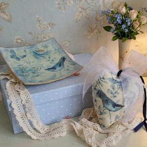 May include: A decorative arrangement on a white table. It includes a blue and white bird-themed heart-shaped item, a square dish, and a polka-dot box. A lamp with a blue lampshade and a vase of flowers are also present.