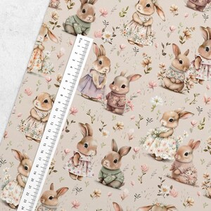 May include: A beige fabric with a repeating pattern of brown bunnies wearing different outfits. The bunnies are surrounded by pink and white flowers. A ruler is visible in the bottom left corner of the image.