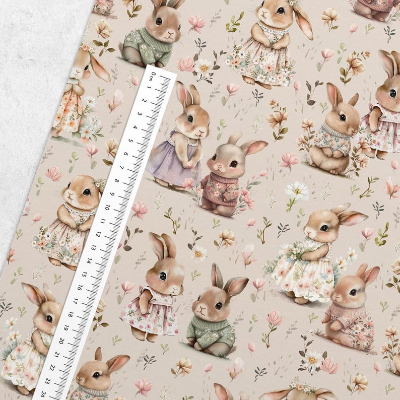 May include: A beige fabric with a repeating pattern of brown bunnies wearing different outfits. The bunnies are surrounded by pink and white flowers. A ruler is visible in the bottom left corner of the image.