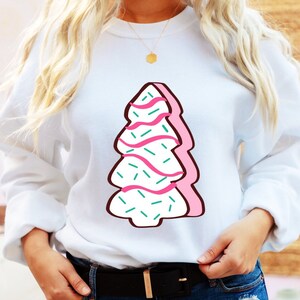 May include: White sweatshirt featuring a pink and green Christmas tree design. The tree is decorated with pink icing and green sprinkles.