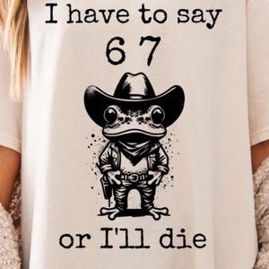 May include: Cream-colored t-shirt with the text "I have to say 67 or I'll die." The shirt features a black illustration of a frog wearing a cowboy hat, a neckerchief, and a gun holster.