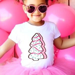 Puede incluir: Una persona joven con una camiseta blanca con un diseño de árbol de jengibre rosa y verde. La persona lleva gafas de sol y un tutú rosa.