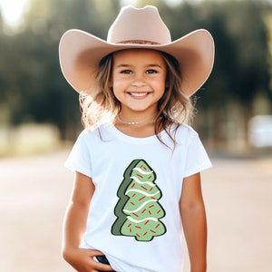 May include: A young girl wearing a white t-shirt with a green Christmas tree design. The tree is decorated with red and white sprinkles. She is also wearing a brown cowboy hat.