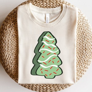 May include: A beige t-shirt featuring a green Christmas tree design. The tree is decorated with white icing and red sprinkles.