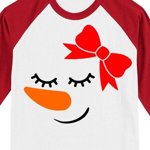May include: A white and red baseball tee shirt with a snowman face design. The snowman has a red bow, closed eyes, an orange nose, and a smiling mouth.