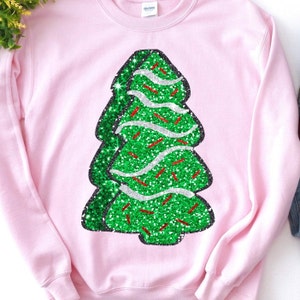 May include: A pale pink sweatshirt with a glittery green Christmas tree design. The tree has white and red accents, and a black outline. The sweatshirt is a Gildan brand.