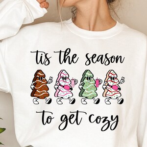 May include: White sweatshirt with the text "Tis the season to get cozy" and four cartoon Christmas trees wearing sunglasses and holding drinks.