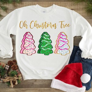 Könnte beinhalten: Weißer Pullover mit dem Text "Oh Christmas Tree" in goldenen Glitzerbuchstaben. Unter dem Text befinden sich drei glitzernde Weihnachtsbaummotive, ein grüner und zwei rosa.
