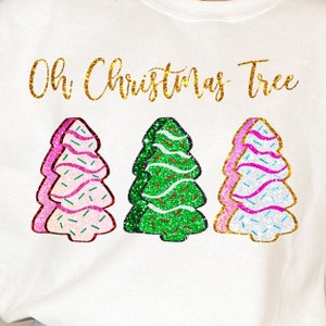 Könnte beinhalten: Weißes T-Shirt mit dem Text "Oh Christmas Tree" in goldenen Glitzerbuchstaben. Unter dem Text befinden sich drei glitzernde Weihnachtsbäume: ein rosa, ein grüner und ein weißer.