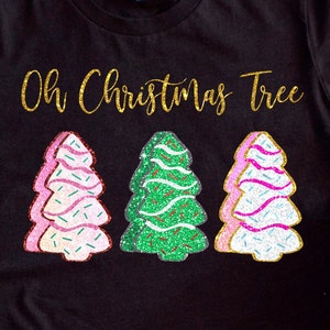 Könnte beinhalten: Ein schwarzes T-Shirt mit goldenen Glitzerbuchstaben, auf dem "Oh Christmas Tree" steht. Unter dem Text sind drei glitzernde Weihnachtsbaumkekse abgebildet. Die Kekse sind rosa, grün und weiß mit Glitzerstreuseln.