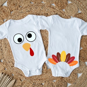 Op de afbeelding: Twee witte baby-rompers met een Thanksgiving-kalkoen-thema. Eén romper heeft een kalkoen gezicht met goggle-ogen, een rode keellap en een oranje snavel. De andere romper heeft een kalkoenstaart met bruine, oranje en rode veren.