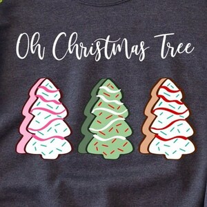 May include: A grey sweatshirt with the text "Oh Christmas Tree" printed in white. Three gingerbread cookie Christmas trees are depicted below the text. The trees are decorated with pink, green, and white icing and sprinkles.