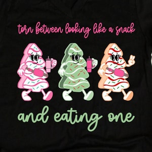 May include: A black t-shirt with the text "torn between looking like a snack and eating one" in pink and green. Three cartoon Christmas trees are walking, each holding a pink drink. The trees are decorated with pink, green, and red frosting.