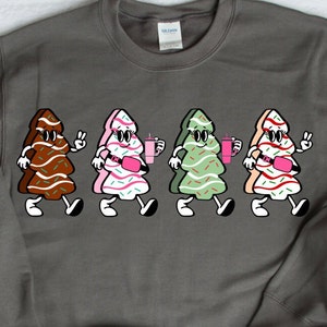 May include: A dark grey sweatshirt with a cartoon design of four walking Christmas trees. Each tree is a different colour, with sprinkles and accessories. The trees are brown, pink, green, and tan, and each is holding a drink.