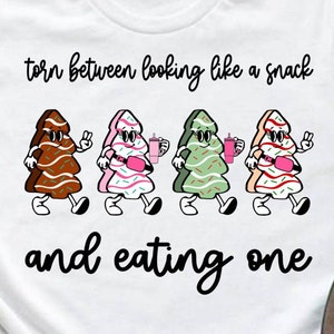 Puede incluir: Una camiseta blanca con el texto "torn between looking like a snack and eating one" impreso en ella. Cuatro árboles de Navidad de dibujos animados caminan por la camiseta, cada uno con una bebida y un cinturón rosa.