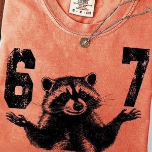 May include: Coral-colored t-shirt featuring a black raccoon graphic with the numbers "6" and "7" on either side. The shirt has a crew neck and a small tag at the top. A silver necklace with a small pendant is draped over the shirt.
