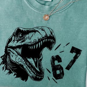 May include: A teal Comfort Colors t-shirt featuring a black graphic of a Tyrannosaurus Rex head with open jaws and the number "67". A silver necklace with a pendant is also visible. The shirt has a crew neck and short sleeves.