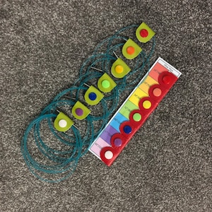 May include: A red leather strip with a colour chart labelled "Yarn Size" and a series of coloured leather loops. The colours are red, orange, yellow, green, blue, purple, and white. The chart shows the yarn size in inches. The leather loops are attached to a series of blue yarn circles.