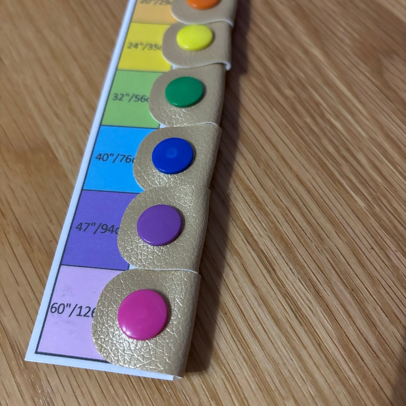 May include: A gold leather measuring tape with a rainbow of colored plastic tabs. The tabs are labeled with measurements in inches and centimeters. The tabs are yellow, green, blue, purple, and pink.