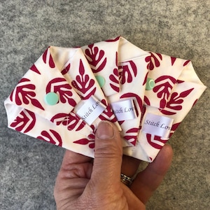 May include: Three white cloth pads with a red leaf pattern and the text "Stitch Love" on each. The pads are folded and held in a hand.