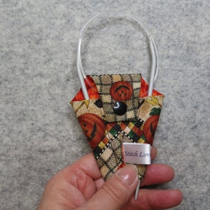 May include: A fabric knitting needle case with a pumpkin and plaid pattern. The case is held by a person's hand. The case has a black button closure and a label that reads "Stitch Love". The case holds two knitting needles.