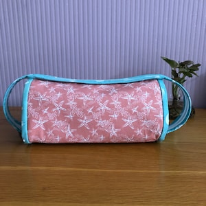 May include: A cylindrical, coral-colored fabric bag with white starfish patterns. The bag has a light blue trim and matching handles. The bag is approximately 10 inches long and 5 inches in diameter, perfect for carrying small items.