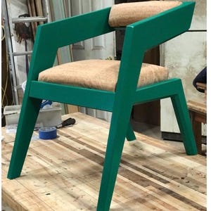May include: A green wooden chair with a light brown fabric seat and back. The chair has a unique angled design and is sitting on a wooden surface.
