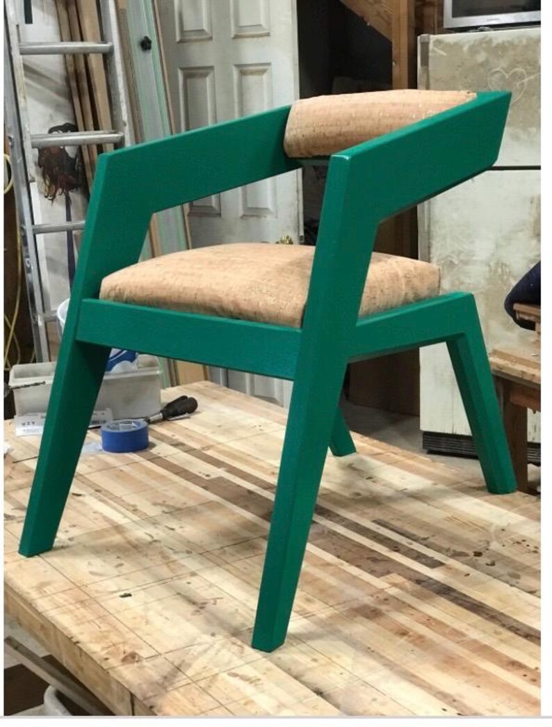 May include: A green wooden chair with a light brown fabric seat and back. The chair has a unique angled design and is sitting on a wooden surface.